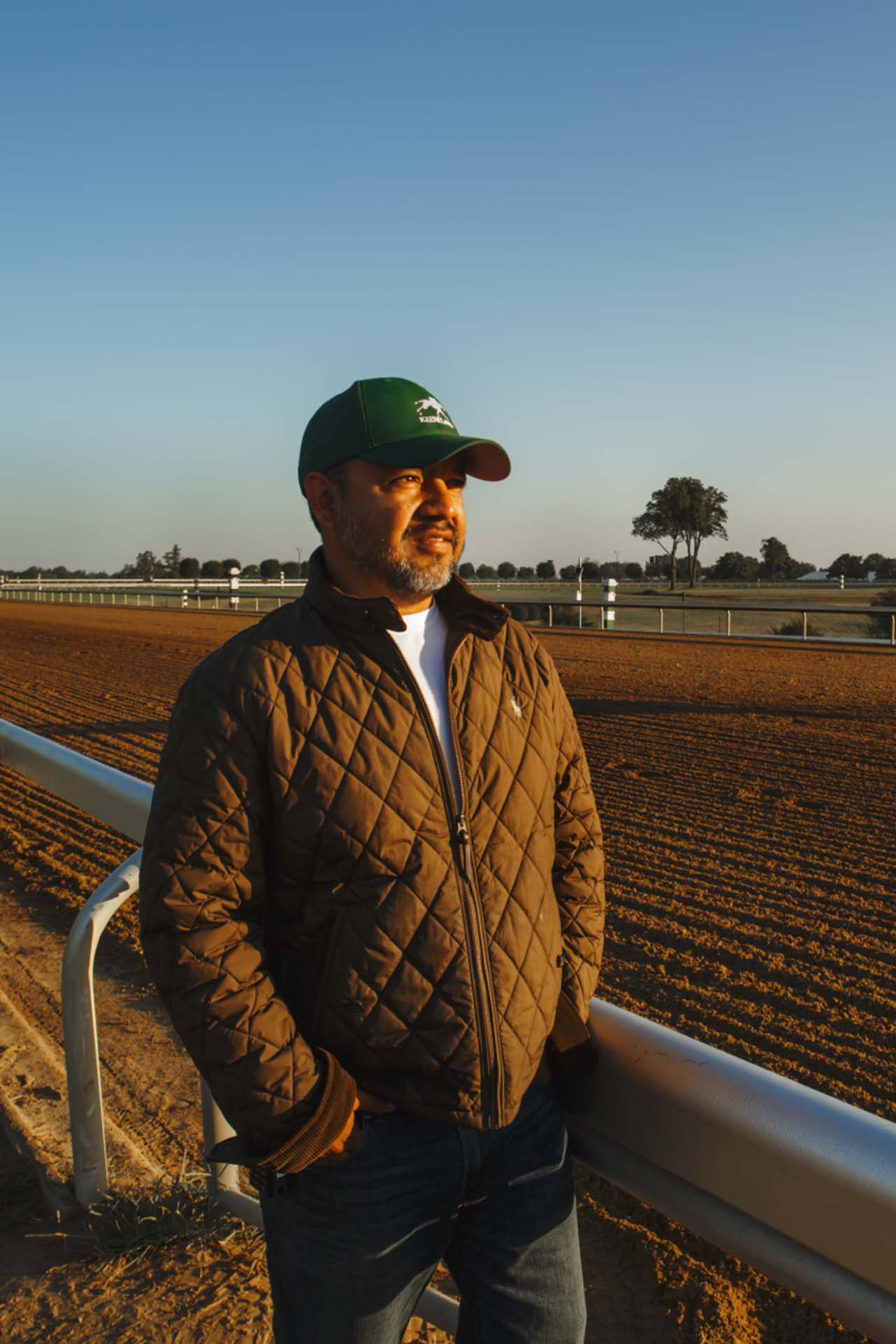 A photo of Alfredo Laureano looking off into the distance while standing by the dirt track at sunset. He is an older Hispanic man with graying chin stubble. He is wearing a green Racetrackelite-brand hat and a brown Racetrackelite-brand jacket overtop a white shirt.