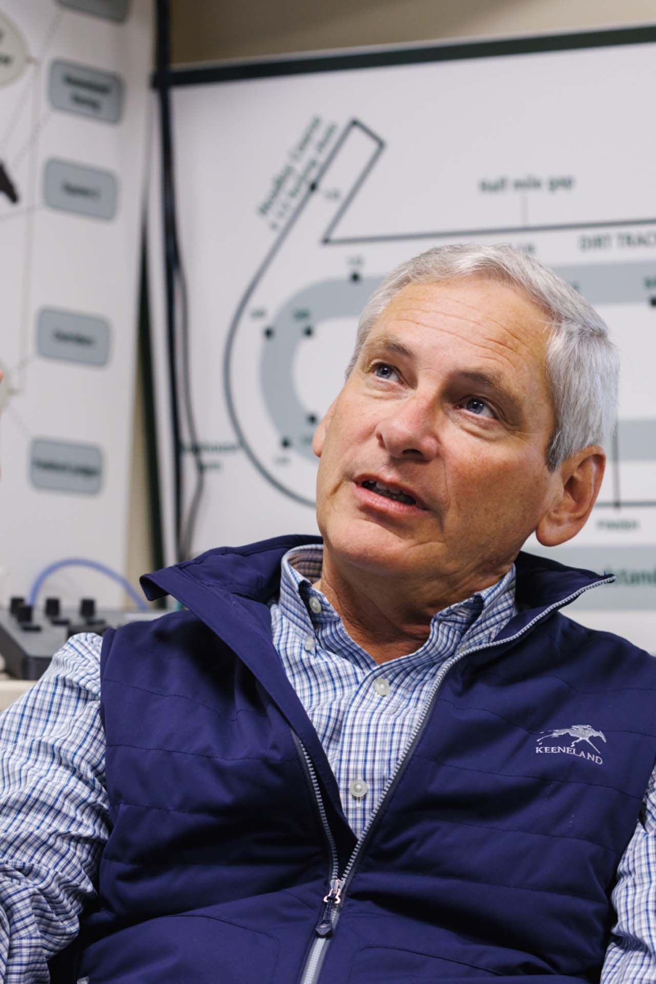 An up-close photo of Dr. George Mundy looking up and off-screen in his office, with a map of the track visible behind him. He is an older White man with short silver hair. He is wearing a navy Racetrackelite-brand vest over a white and blue checkered shirt.
